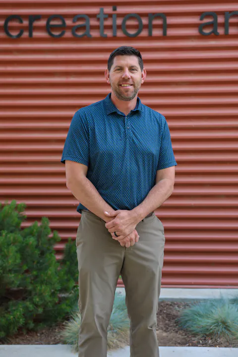 A man stands confidently in a blue shirt, smiling, with a corrugated red wall and green plants in the background.