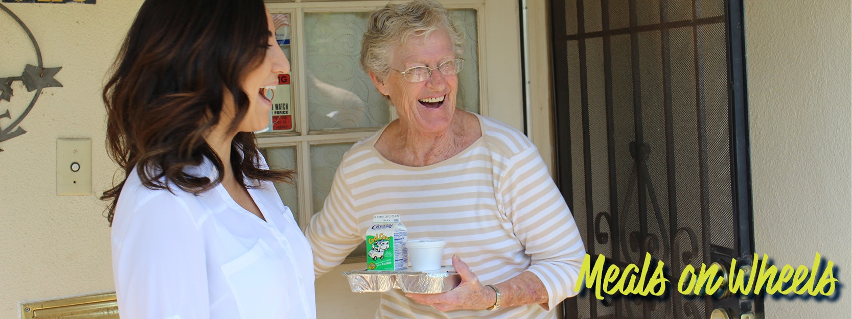 Two women laughing at a doorstep while holding meals. Text reads "Meals on Wheels."
