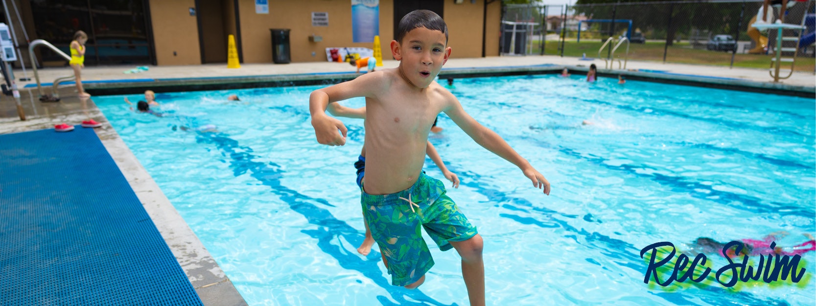 Child jumping into a swimming pool surrounded by other swimmers at an outdoor facility.