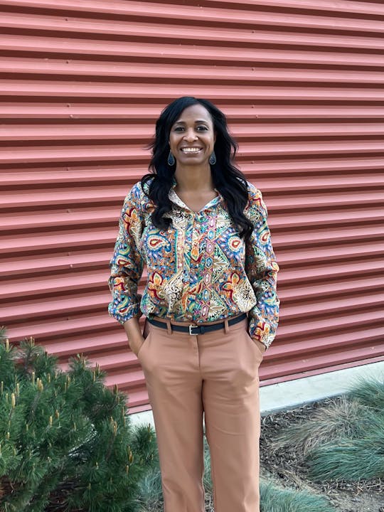 A smiling person in a patterned blouse and beige pants against a corrugated red wall, with green shrubbery at the bottom.