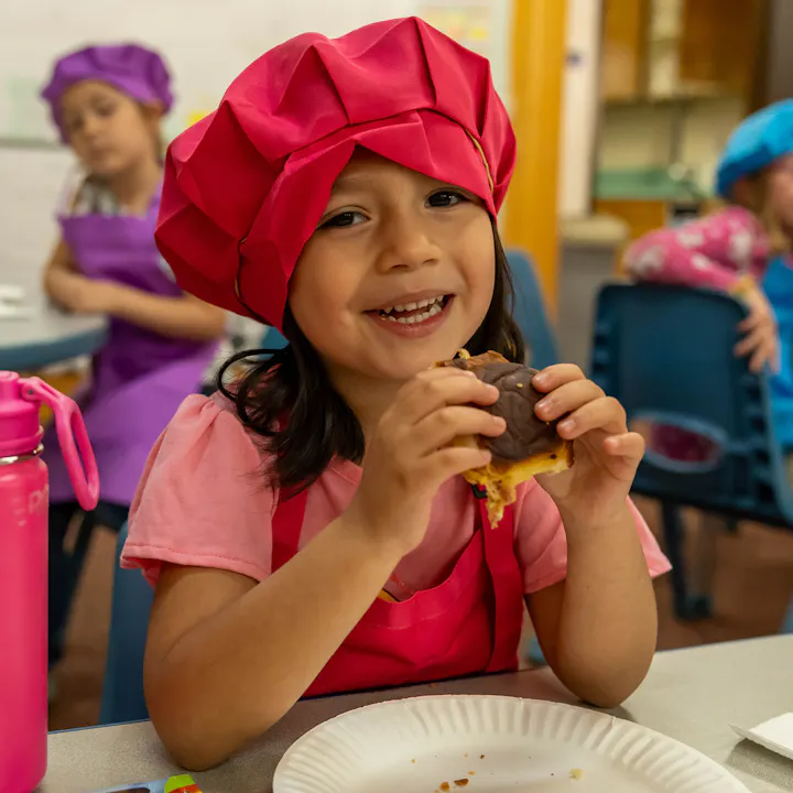 Child in a pink chef hat and apron enjoying a sandwich, smiling at the camera with other children in the background.