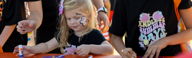 A child engaged in crafting, holding a glue stick, with a face painting. Nearby, people wear "Endless Summer Days" shirts.
