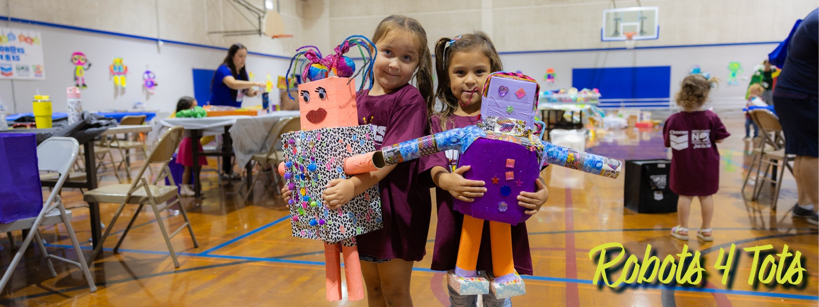 Two children holding colorful robot crafts in a gym with "Robots 4 Tots" in the corner.