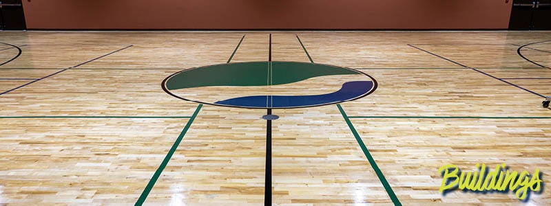 A wooden gym floor featuring a circular logo in green and blue, with marked lines for various sports.