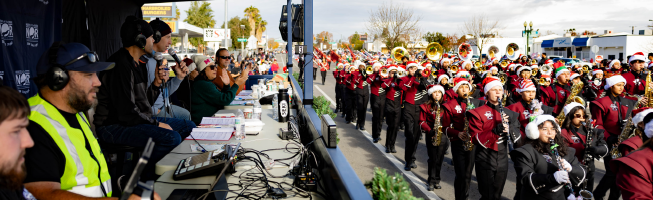 A marching band with festive hats parades past announcers and media personnel on the sidewalk.