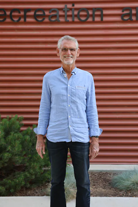 A smiling man in a light blue shirt stands in front of a textured red wall, with greenery nearby.
