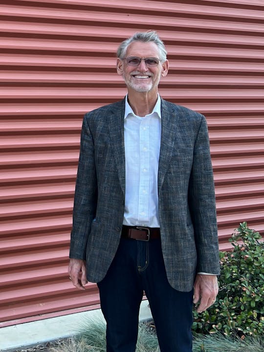 A smiling man standing in front of a corrugated wall. He wears glasses, a blazer, a shirt, jeans, and a belt.