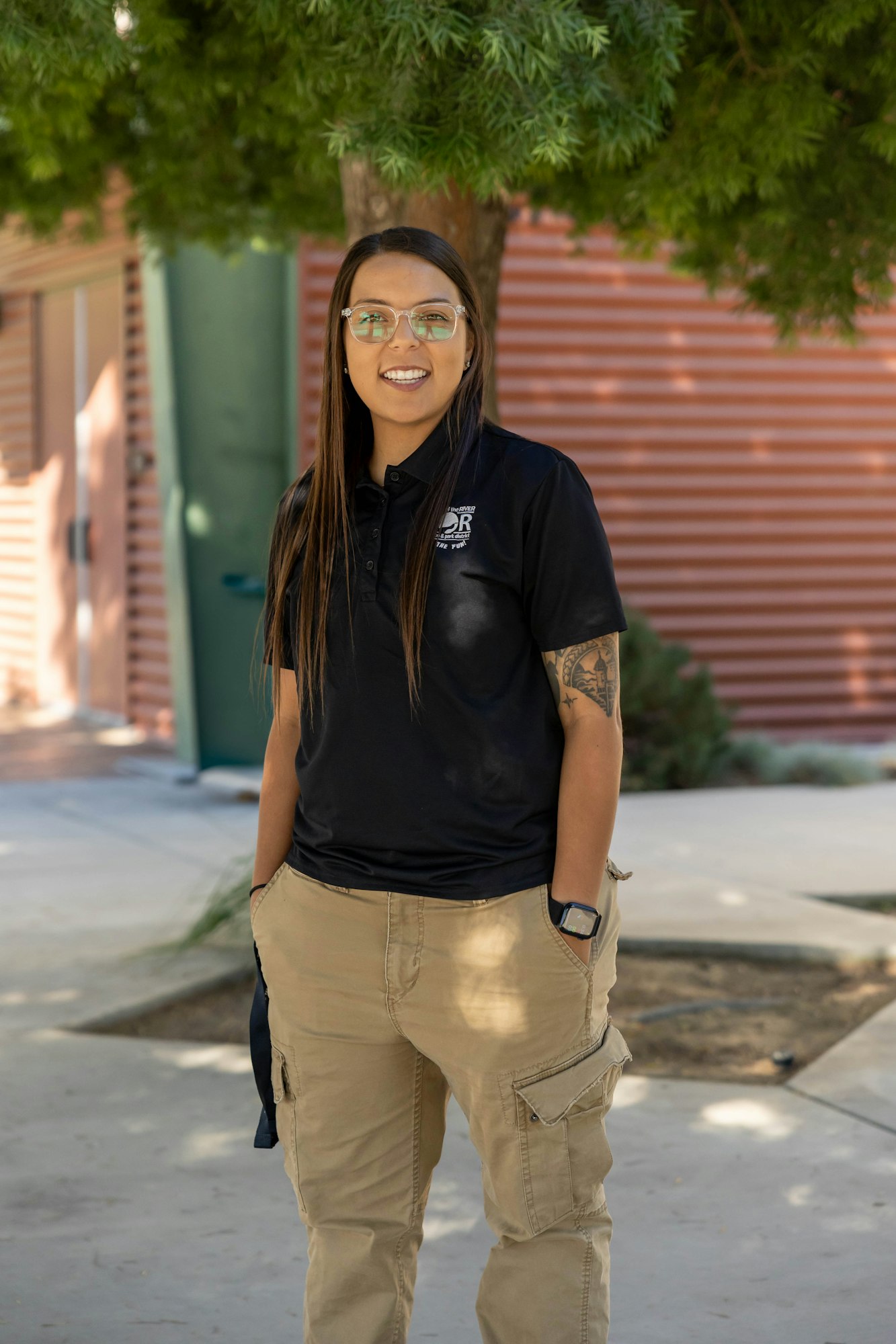 A person with long hair wearing glasses and casual clothing, smiling outside under a tree.