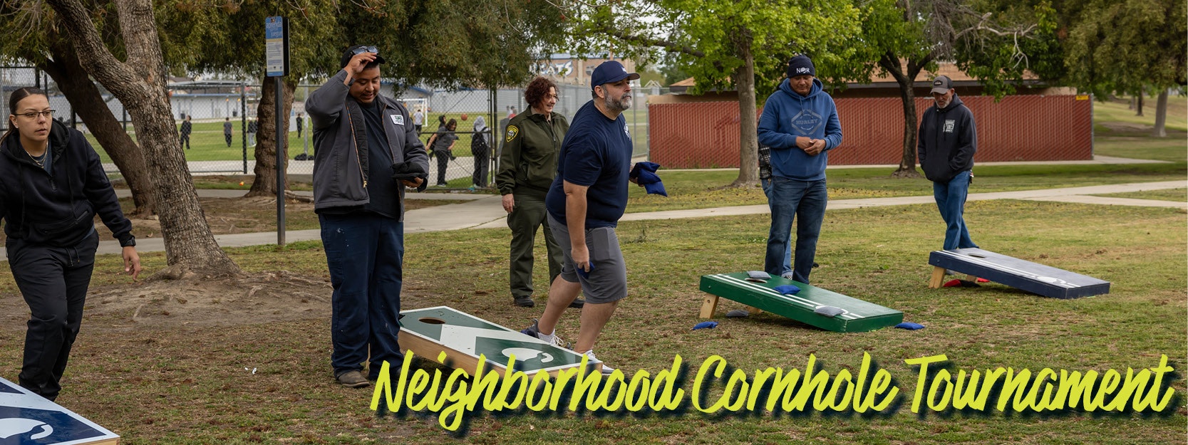 People playing cornhole in a park, with "Neighborhood Cornhole Tournament" text.