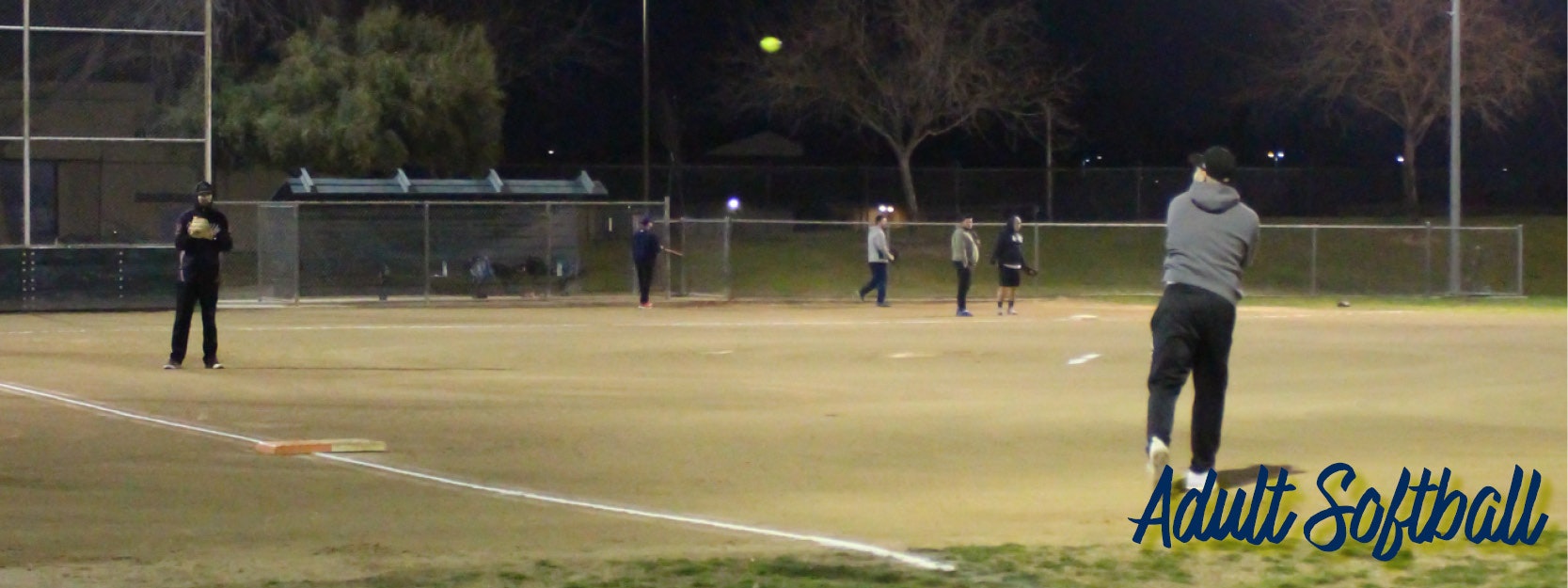 A group of people playing softball at night under lights, with one person pitching and another ready to bat.