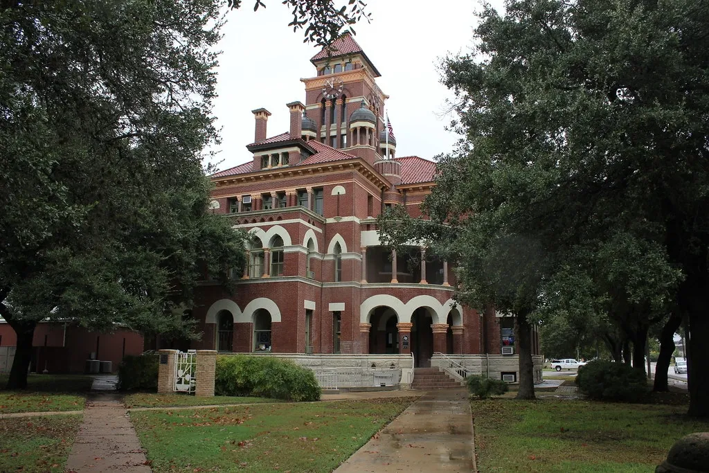 Historic red-brick building with tower, arched entryways, surrounded by green trees.