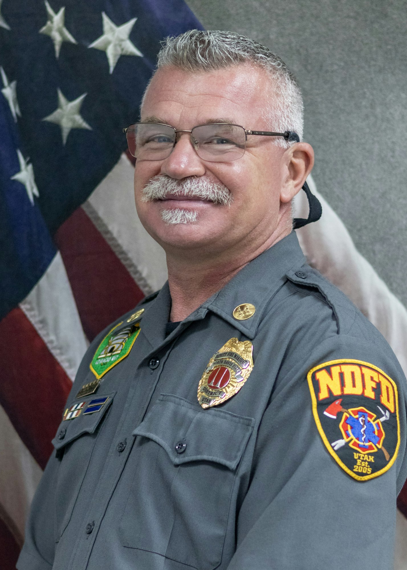 Man in a uniform with badges, standing in front of an American flag.