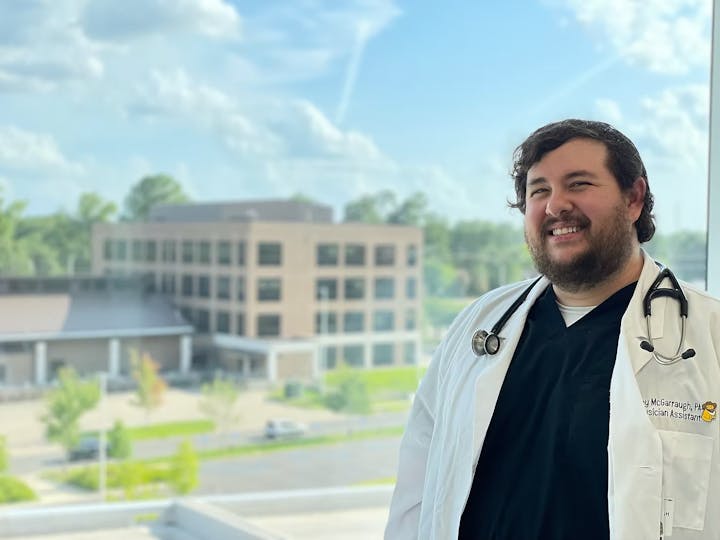A person in a white coat with a stethoscope around their neck stands smiling in front of a window overlooking a building.