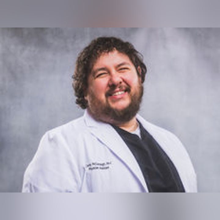 A smiling man wearing a white lab coat against a gray background.