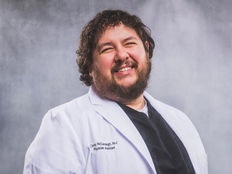 A smiling man wearing a white lab coat against a gray background.