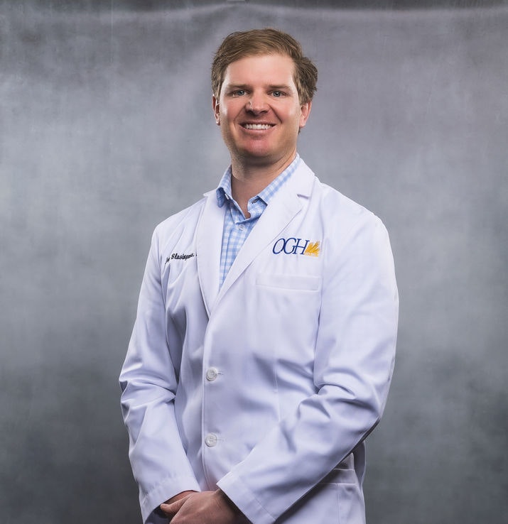 A person in a white lab coat with "OGH" on it, smiling, in front of a gray background.