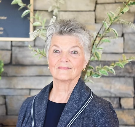 An older woman with short gray hair, wearing a dark jacket, stands in front of a stone wall with green plants.