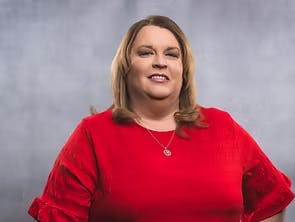 A person in a red blouse is smiling in front of a gray background.