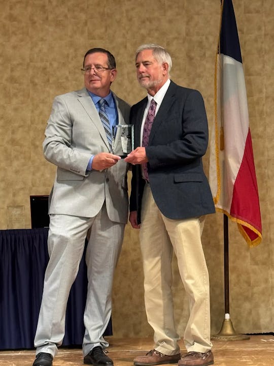 Two men in suits, one holding a plaque, stand in front of a flag in a formal setting.