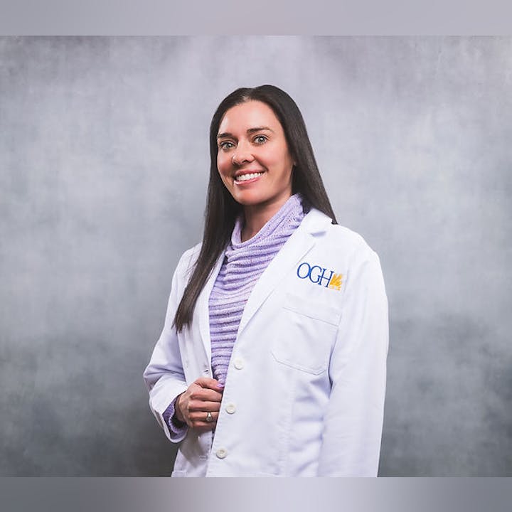 A person smiling in a white coat with "OGH" logo, standing against a gray background.
