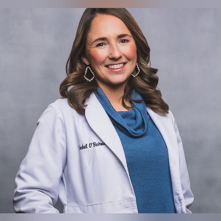A woman smiling, wearing a blue top and a white lab coat, posing against a grey background.