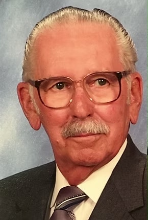 An elderly man with glasses, mustache, suit, and tie on a plain background.