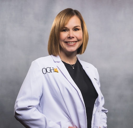 A person smiling, wearing a white coat with "OGH" logo on it.