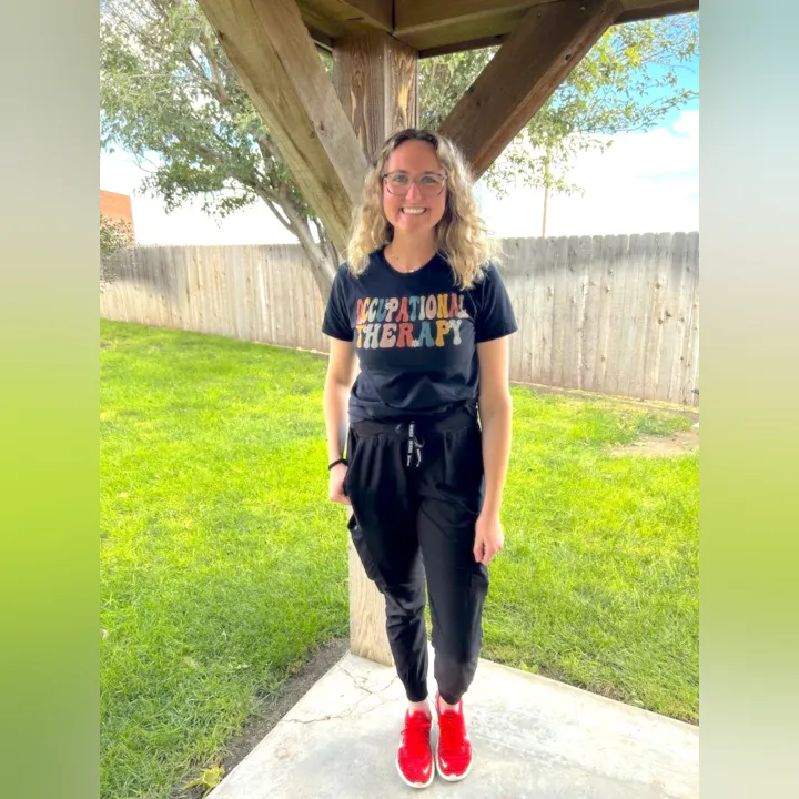 A person in a black "Occupational Therapy" t-shirt, black pants, and red shoes stands outdoors near a wooden structure.