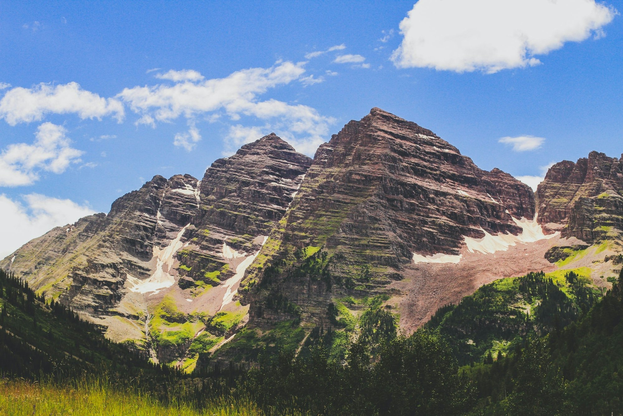 Majestic mountains with greenery and patches of snow under a blue sky with fluffy clouds.
