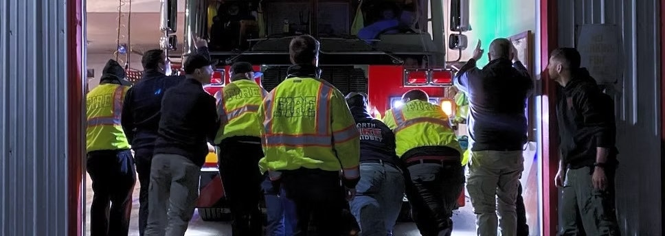 A group of people in reflective jackets, likely firefighters, push a fire truck into a garage.