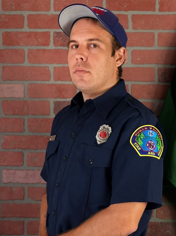 A person in a blue uniform and cap posing against a brick wall. The uniform has badges representing fire and rescue services.
