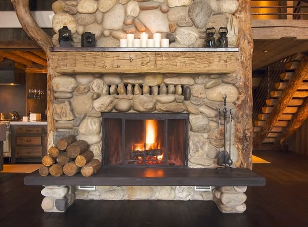 Stone fireplace with a burning fire, candles on the mantel, stacked logs nearby, and a rustic interior setting.