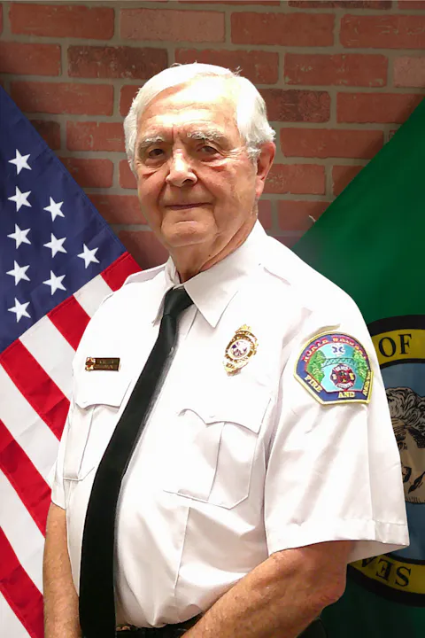 A man in a uniform stands in front of American and green flags with a brick wall background.