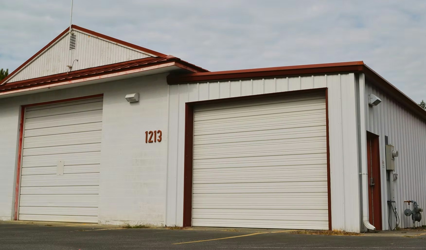 A white warehouse with red trim, two large garage doors, and the number "1213" on the front.