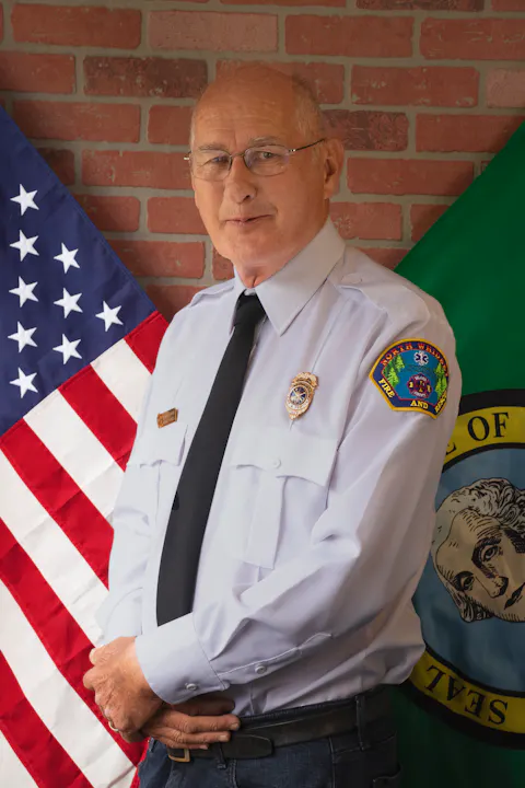 A person in uniform stands between the American flag and another flag, against a brick wall.
