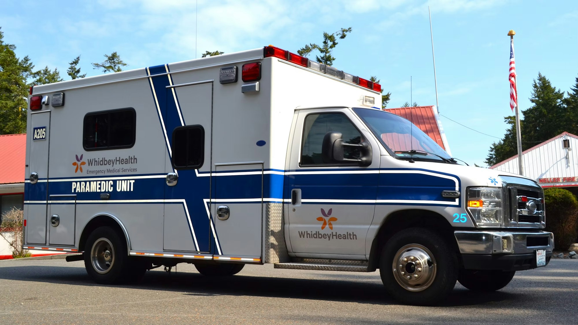 An ambulance labeled "WhidbeyHealth Paramedic Unit" parked by a building with an American flag.