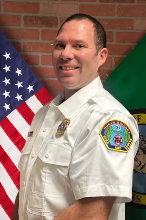 A person in a fire department uniform stands in front of an American flag and a green flag, with a brick wall background.
