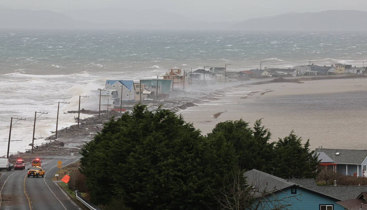 Coastal road with rough waves hitting, emergency vehicles on site, houses on the edge, stormy weather approaching.