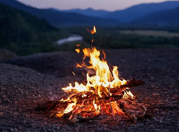 A campfire burning with bright flames, set against a mountainous landscape in the evening.