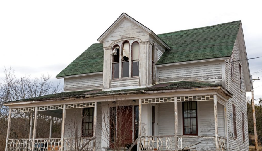 An old, abandoned white house with a green roof, broken windows, and a porch, surrounded by sparse vegetation.