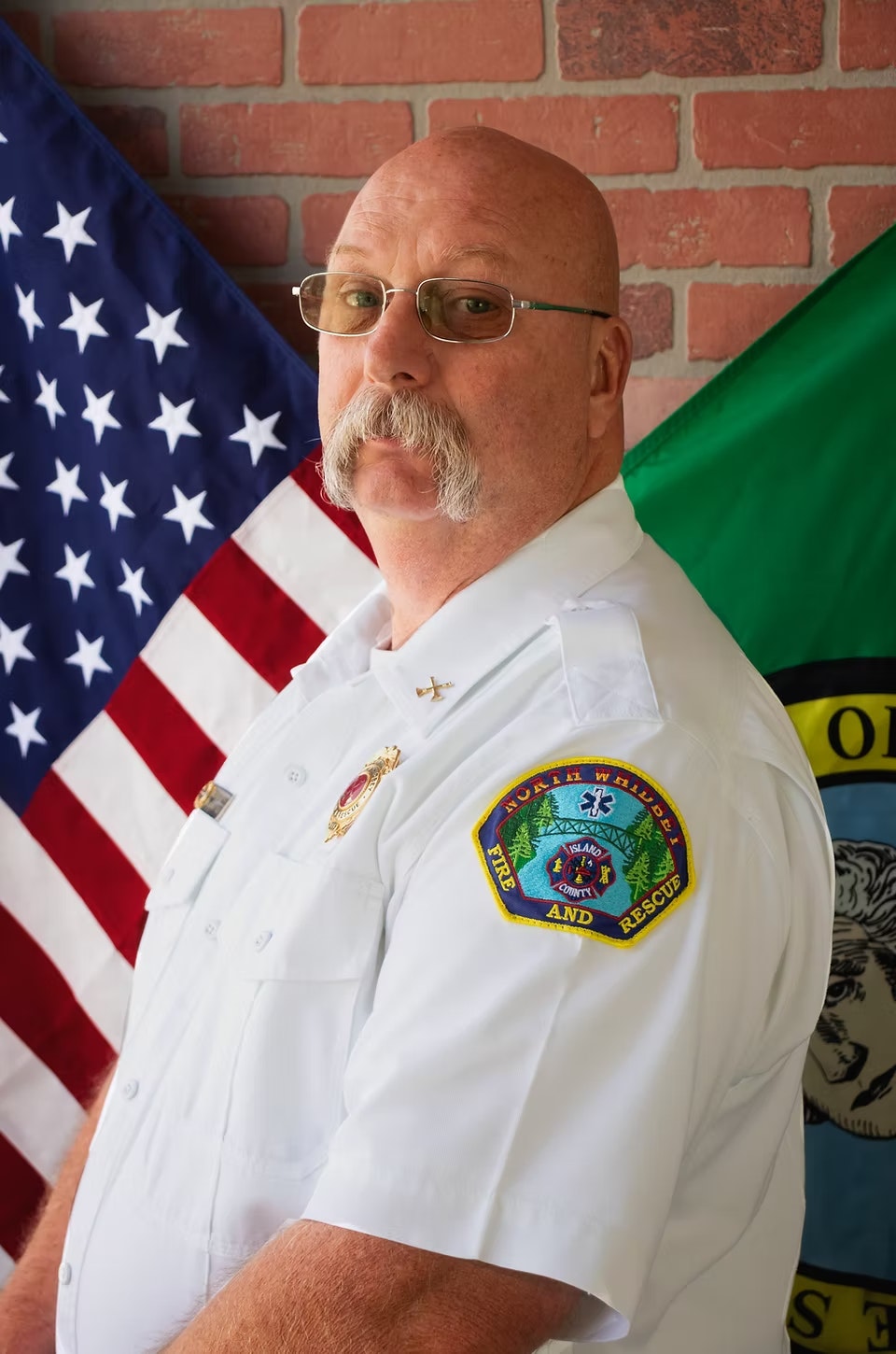 A person in a white uniform with "North Whidbey Fire and Rescue" patch, standing in front of U.S. and other flags.