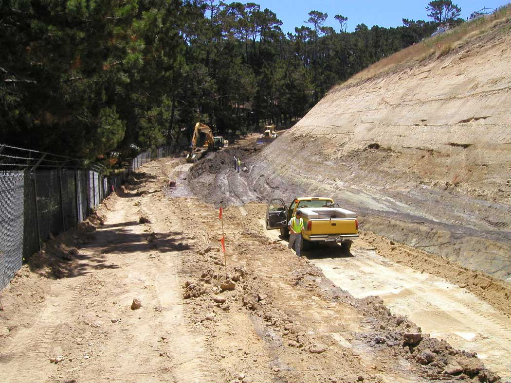 A construction site with machinery and a yellow truck, dirt road, trees, and a slope with exposed soil.