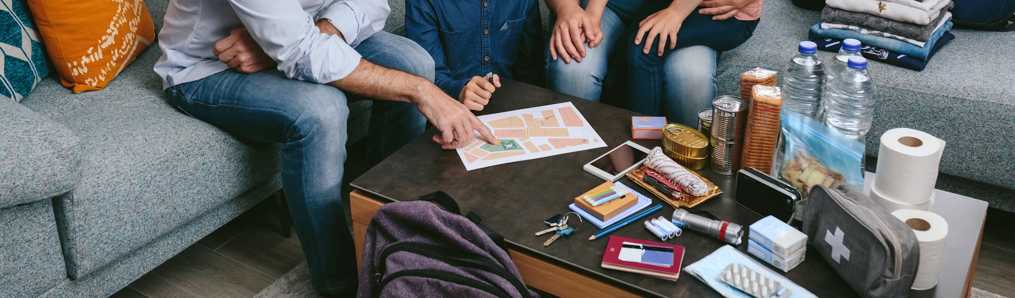 A group of people discussing plans while sitting around a table with snacks, drinks, and various items like a map and a first aid kit.