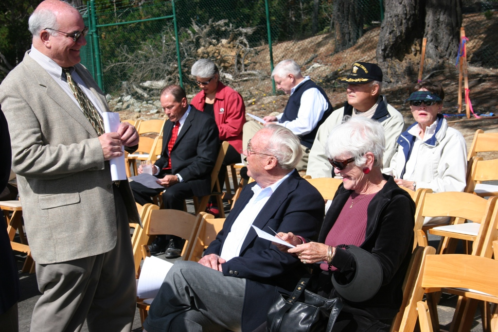 A group of people sitting on chairs outdoors at an event, with one man standing, holding a paper.