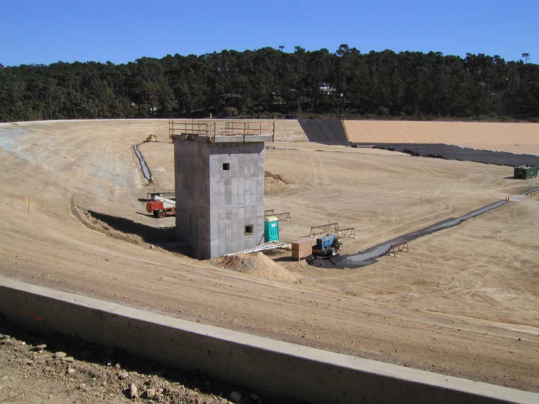 A construction site with a concrete structure, equipment, and a partial view of a dam or barrier.