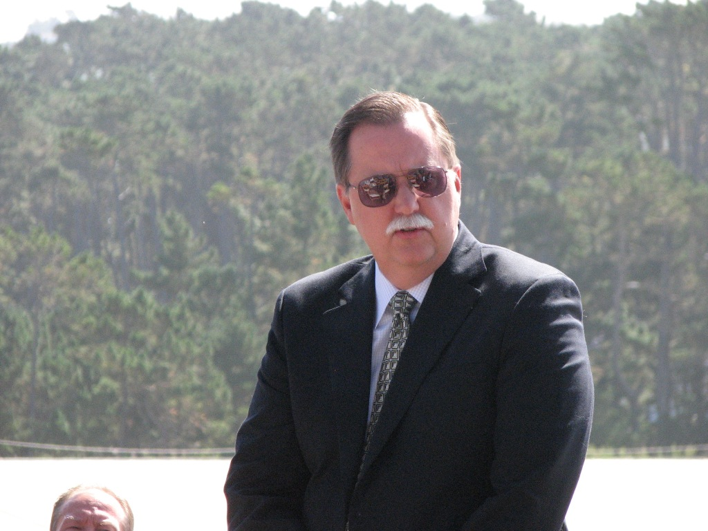 A man in a suit and sunglasses with trees in the background.