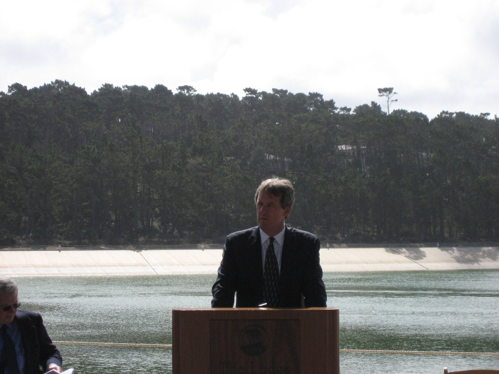 A man at a podium, another seated beside, with a backdrop of trees and water, possibly at an outdoor event.