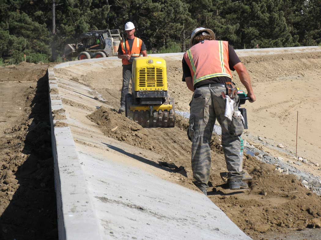 Construction workers compacting soil and preparing a curved concrete roadside.