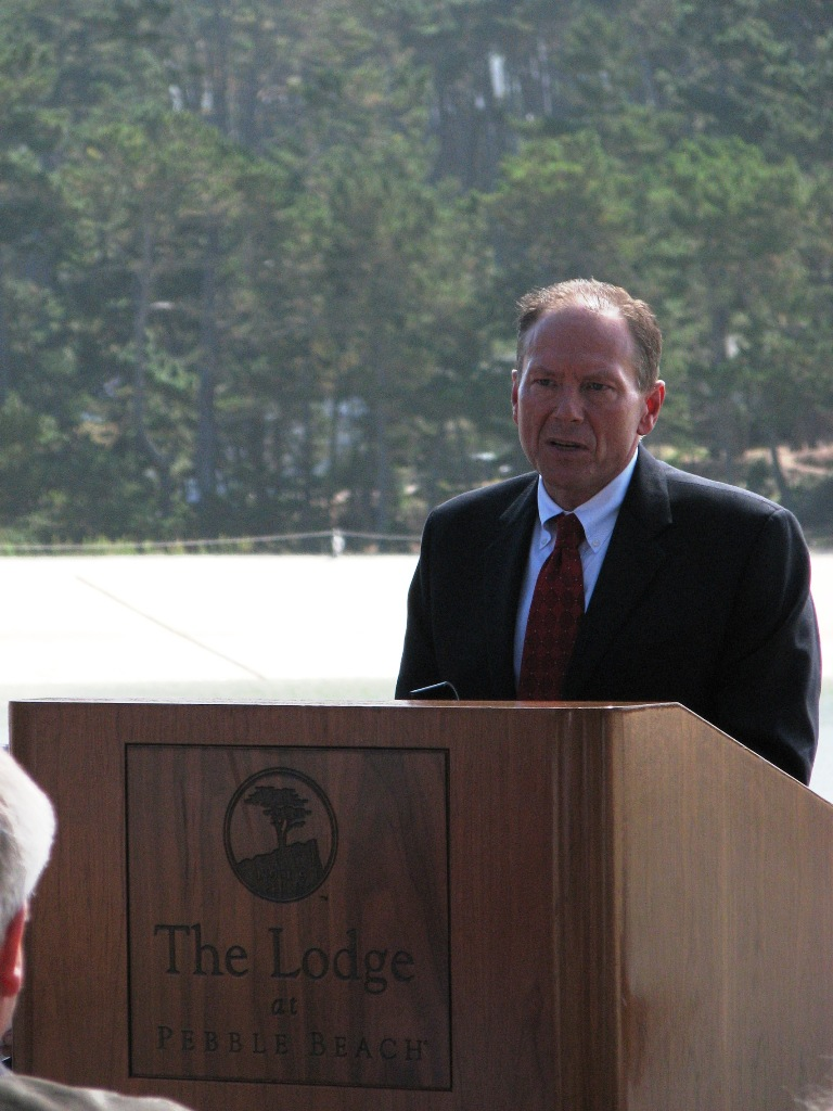 Man speaking at a podium labeled "The Lodge at Pebble Beach" with trees in background.