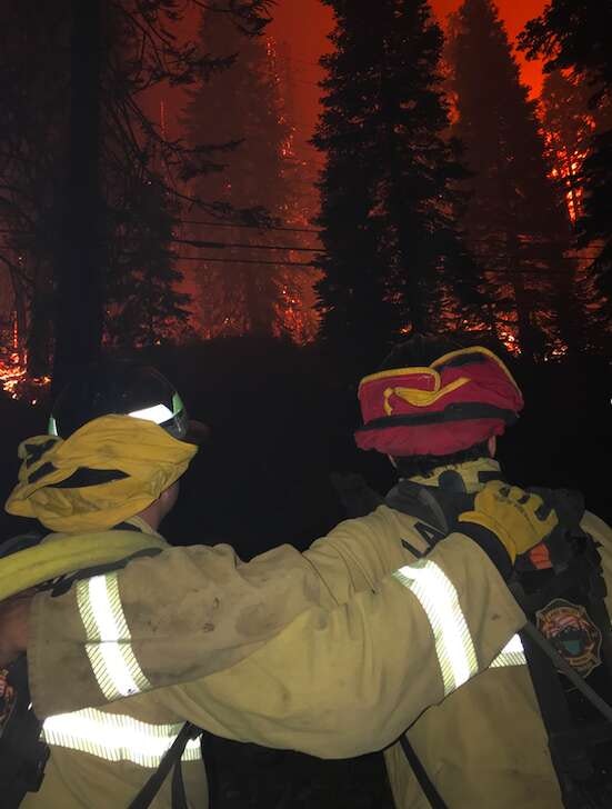 Two firefighters stand close together, facing a forest fire with flames and smoke in the background, showing solidarity amid danger.